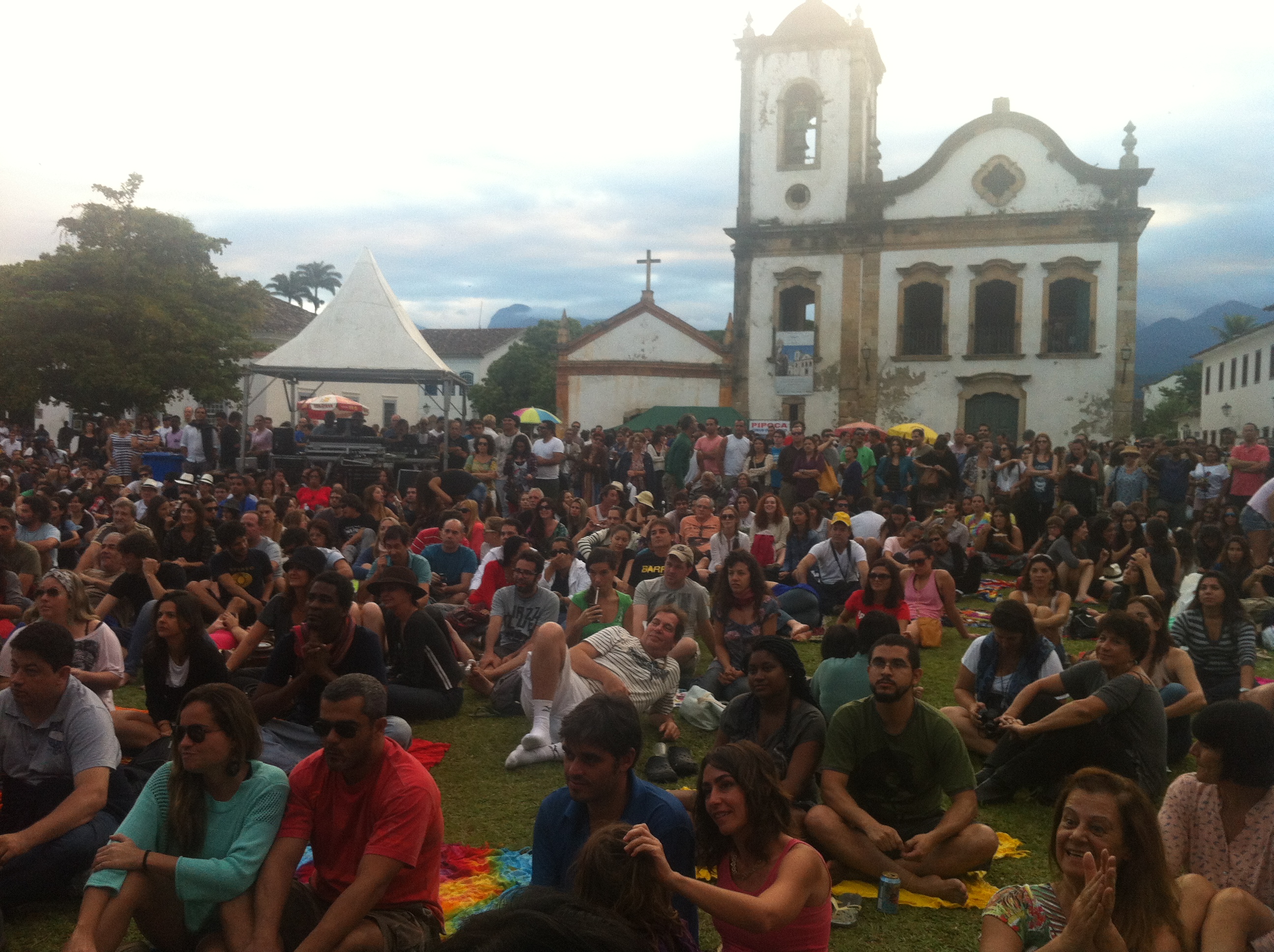 Público sentado no gramado em frente ao palco durante shows da tarde em frente à Igreja de Santa Rita. Muita gente sentada, num clima muito gostoso, público aplaudindo